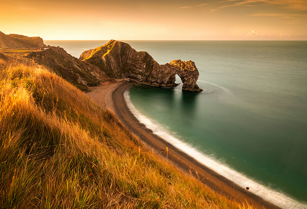 A sweeping golden-toned coastline leads to a sea arch over the ocean in front of a setting sun.